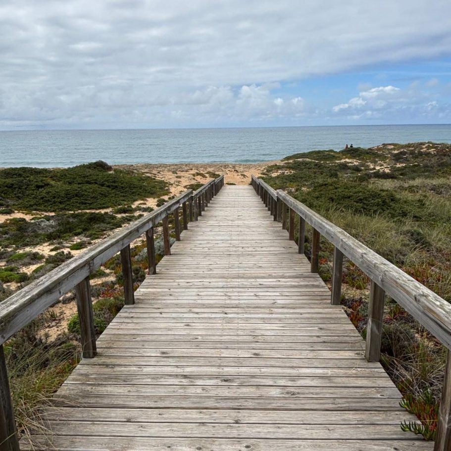 Steg in Brückenform mit Geländer, der durch Steilküstenvegetation zum Strand runter führt, Himmel reisst gerade auf